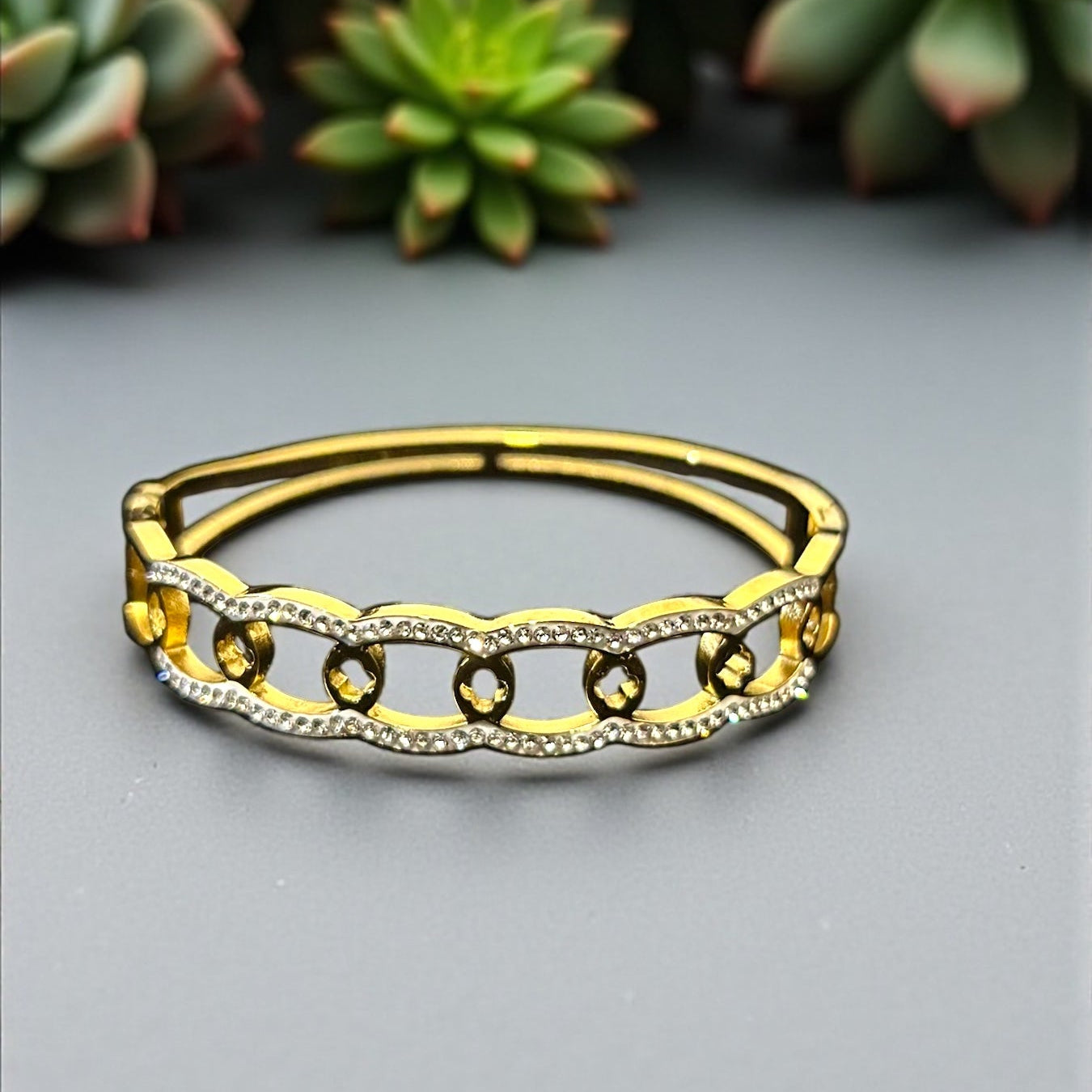 Gold bracelet with intricate design on a gray background with succulents